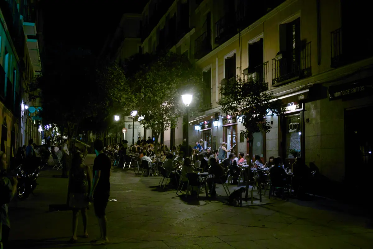 A narrow Malasaña street at night, with people eating at café tables under street lamps.